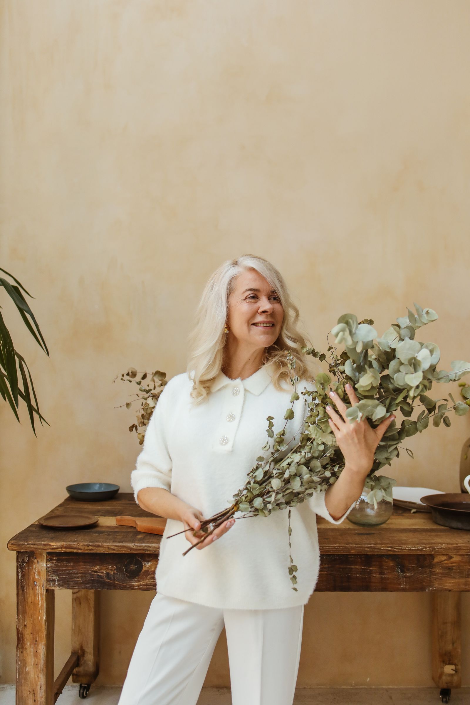 A smiling older woman holding eucalyptus branches in a calm, bright room at home